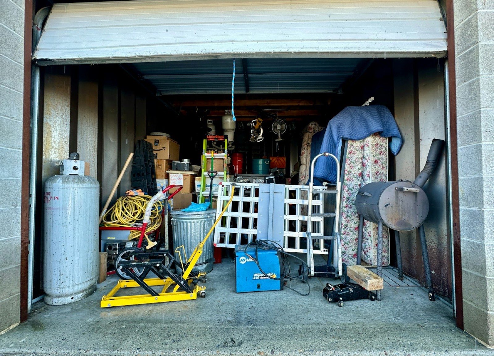 A storage unit packed to the roof with a mattress, ladders, a welder, a propane tank and stacked boxes, before clearing.